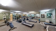 Located at 2150 Wilson Ave in Saint Paul, Minnesota, this fitness center offers a practical and well-organized space for exercise and wellness. The facility occupies a single floor and is divided into two functional areas: a spacious exercise room and a dedicated bathroom.

The exercise room is the heart of this facility, generously sized to accommodate a wide variety of cardiovascular and strength-training equipment. The room features multiple treadmills, elliptical machines, a stationary bike, cable weight machines, a weight bench with free weights, and a dumbbell rack. Large wall-to-wall mirrors line one side of the room, enhancing the sense of openness and allowing users to monitor their form during workouts. The space is finished with durable carpet flooring, a textured drop ceiling with recessed fluorescent lighting, and soft sage-green walls. Fitness reference posters and a posted set of fitness center rules are displayed for user guidance.

The adjoining bathroom is designed with accessibility in mind, featuring tiled walls in neutral earth tones, tile flooring, and grab bars for added safety. The space includes a toilet, a wall-mounted sink with a vanity mirror, and a storage cabinet, providing all necessary amenities for post-workout freshening up. Together, these two rooms create a complete and functional fitness environment.