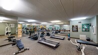 Located at 2150 Wilson Ave in Saint Paul, Minnesota, this fitness center offers a practical and well-organized space for exercise and wellness. The facility occupies a single floor and is divided into two functional areas: a spacious exercise room and a dedicated bathroom.

The exercise room is the heart of this facility, generously sized to accommodate a wide variety of cardiovascular and strength-training equipment. The room features multiple treadmills, elliptical machines, a stationary bike, cable weight machines, a weight bench with free weights, and a dumbbell rack. Large wall-to-wall mirrors line one side of the room, enhancing the sense of openness and allowing users to monitor their form during workouts. The space is finished with durable carpet flooring, a textured drop ceiling with recessed fluorescent lighting, and soft sage-green walls. Fitness reference posters and a posted set of fitness center rules are displayed for user guidance.

The adjoining bathroom is designed with accessibility in mind, featuring tiled walls in neutral earth tones, tile flooring, and grab bars for added safety. The space includes a toilet, a wall-mounted sink with a vanity mirror, and a storage cabinet, providing all necessary amenities for post-workout freshening up. Together, these two rooms create a complete and functional fitness environment.
