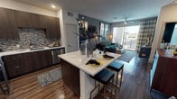 This stylish single-floor unit showcases a thoughtfully designed open-concept layout that seamlessly connects the kitchen, dining, and living areas. The kitchen is outfitted with dark wood cabinetry, a textured stone tile backsplash, and stainless steel appliances, including a dishwasher. A large central island with a bright white countertop serves as both a prep space and a casual dining area, accommodating bar-height seating with sleek black and gold stools. Rich hardwood flooring flows throughout the entire unit, adding warmth and cohesion to the space. The living area is anchored by a ceiling fan and features floor-to-ceiling sliding glass doors that open to a private balcony, flooding the interior with abundant natural light. Neutral and deep teal accent walls provide a modern, sophisticated backdrop that complements the curated décor. The living room comfortably accommodates a full sofa and coffee table, and is further enhanced by large framed wall art and a stylish floor lamp. The balcony, accessible through the sliding glass doors and dressed with patterned drapes, extends the living space outdoors. Overall, the unit delivers a contemporary urban aesthetic with high-end finishes and a bright, open ambiance.