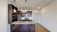 This well-appointed one-bedroom, one-bathroom unit is located in Jenks, Oklahoma, offering a thoughtfully designed single-level layout. The open-concept kitchen and living area serves as the heart of the space, anchored by a substantial granite-topped island with an undermount sink, providing both prep space and casual seating. Rich, dark-stained cabinetry lines the kitchen walls alongside a full suite of stainless steel appliances, including a refrigerator, electric range, and built-in microwave. A white subway tile backsplash adds a clean, modern contrast, while pendant lighting and a track light fixture above illuminate the space with a contemporary industrial flair. An open shelving unit adds additional display and storage options. The living area benefits from large windows flanking the front entry door, allowing natural light to fill the room. Warm wood-look plank flooring flows consistently throughout the unit, creating a cohesive and inviting atmosphere. The bedroom is a comfortable, well-proportioned room featuring a ceiling fan and closet access. The bathroom includes a full tub-shower combination with a curved curtain rod, a toilet, and an overhead storage cabinet with matching dark-stained cabinetry. Clean white walls and neutral finishes throughout the unit create a bright, move-in-ready environment.