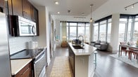 This contemporary single-floor space showcases a thoughtfully designed open-concept layout that seamlessly connects the kitchen, dining, and living areas. The kitchen is appointed with dark-toned cabinetry, stainless steel appliances including a built-in microwave and range, and a large center island topped with a crisp white countertop and an undermount sink. Pendant lighting hangs above the island, adding a refined, modern touch. A textured area rug grounds the kitchen corridor, adding warmth against the rich hardwood flooring that runs throughout the space. Beyond the kitchen, the living area is bathed in abundant natural light courtesy of expansive floor-to-ceiling windows that wrap around the space, offering sweeping urban views. The living area is furnished with contemporary seating and accented by soft, neutral tones that create a calm and inviting atmosphere. Track lighting and recessed ceiling fixtures provide flexible illumination throughout. Sheer curtains frame the windows, softening the transition between interior comfort and the city backdrop. The overall ambiance is clean, bright, and modern — a polished urban living environment that balances style with functionality.