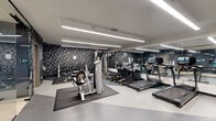 Located at 207 S Maple St in Mount Prospect, IL, this fitness center offers a thoughtfully designed, single-floor space divided into two functional areas. The main gym floor is defined by a striking black geometric accent wall that creates a bold visual backdrop against the clean white ceiling, from which long linear LED light fixtures provide bright, even illumination throughout. Floor-to-ceiling mirrors line one wall, visually expanding the space and allowing users to monitor their form. The main area is well-stocked with a row of treadmills and elliptical machines, a cable strength machine, a seated row machine, and a full dumbbell rack, all arranged across protective rubber floor mats. A water station is also conveniently accessible. A separate, quieter studio room features muted gray walls, wood-look flooring, protective matting, a stationary bike, and a wall-mounted interactive fitness mirror, making it ideal for focused cycling sessions or guided workouts. The dual-zone layout effectively separates cardio and strength training from private, low-impact exercise. Overall, the space maintains a modern, professional aesthetic with a monochromatic color palette, efficient use of floor space, and high-quality equipment that supports a comprehensive fitness experience.