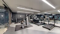 Located at 207 S Maple St in Mount Prospect, IL, this fitness center offers a thoughtfully designed, single-floor space divided into two functional areas. The main gym floor is defined by a striking black geometric accent wall that creates a bold visual backdrop against the clean white ceiling, from which long linear LED light fixtures provide bright, even illumination throughout. Floor-to-ceiling mirrors line one wall, visually expanding the space and allowing users to monitor their form. The main area is well-stocked with a row of treadmills and elliptical machines, a cable strength machine, a seated row machine, and a full dumbbell rack, all arranged across protective rubber floor mats. A water station is also conveniently accessible. A separate, quieter studio room features muted gray walls, wood-look flooring, protective matting, a stationary bike, and a wall-mounted interactive fitness mirror, making it ideal for focused cycling sessions or guided workouts. The dual-zone layout effectively separates cardio and strength training from private, low-impact exercise. Overall, the space maintains a modern, professional aesthetic with a monochromatic color palette, efficient use of floor space, and high-quality equipment that supports a comprehensive fitness experience.
