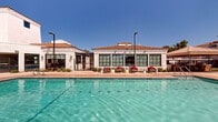 This expansive outdoor amenity space showcases a resort-style design rooted in Spanish Colonial architecture. The centerpiece is a large, crystal-clear swimming pool with decorative water features that arc gracefully into the water, creating a lively and inviting atmosphere. Adjacent to the main pool sits a bubbling spa, offering a relaxing retreat separate from the primary swimming area. The surrounding deck is finished in warm-toned pavers and is generously appointed with lounge chairs and seating clusters accented with bold red cushions, providing ample space for sunbathing and socializing. A pergola-shaded lounge area and market umbrellas offer welcome relief from the sun. The bordering clubhouse building features white stucco exteriors, terracotta tile rooflines, and large multi-pane windows that flood interior spaces with natural light — hallmarks of the Spanish Colonial style. Tall palm trees and mature landscaping frame the space, adding to the warm, desert-resort ambiance. The pool deck is enclosed by a sleek iron fence, ensuring privacy and security throughout the area. Under open, vivid blue skies, the entire space exudes a relaxed yet polished aesthetic, making it an ideal setting for both leisure and community gathering.