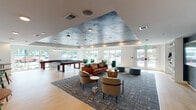 This single-floor game room in Mary Esther, Florida, is a well-appointed recreational space designed for both entertainment and relaxed socializing. The room's most striking architectural feature is its tray ceiling, finished with a dramatic blue-gray textured treatment that adds depth and visual interest overhead. Light wood-look flooring runs throughout, creating a warm, clean foundation that complements the contemporary décor.

The gaming area is anchored by a full-size pool table and a shuffleboard table, both positioned near large windows that allow abundant natural light to fill the space. These expansive windows also offer open views of the surrounding exterior, enhancing the airy atmosphere. Nearby, emerald green accent chairs and bar-height bistro tables provide casual seating for spectators or those looking to unwind.

A dedicated lounge area features a rich cognac leather sofa, rounded accent chairs, and a patterned area rug, all centered around a built-in media wall with a large flat-screen television, open shelving, and bold teal accent panels. Abstract artwork adds a modern artistic touch to this section of the room.

A separate nook includes high-top bistro tables and wood-and-metal bar stools set against white subway tile wainscoting, providing a casual spot for conversation. Recessed lighting throughout ensures the entire space is consistently well-lit and inviting.