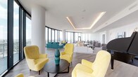 This elegant high-rise amenity space is designed to impress, offering an open, single-floor layout bathed in natural light from sweeping floor-to-ceiling curved glass windows that frame breathtaking panoramic city skyline views. The interior is anchored by a crisp, all-white palette complemented by light hardwood flooring throughout, creating a bright and airy atmosphere.

The expansive lounge area is thoughtfully divided into multiple seating zones, including clusters of vibrant yellow velvet armchairs arranged around round coffee tables, a teal sectional sofa seating area positioned directly against the curved window wall to maximize views, and a more formal lounge zone featuring plush white sectional sofas and gray accent chairs. Tray ceilings with warm recessed LED cove lighting add architectural depth and a refined glow to the space.

A standout feature is the grand black piano, positioned as a focal point alongside curated contemporary artwork displayed on wainscoted white walls, flanked by gold wall sconces. A flat-screen television is also integrated into this area, making it functional for events and gatherings.

The formal dining area accommodates a large party at a sleek dark dining table with modern black-frame chairs, illuminated by a striking cluster of geometric gold pendant lights. White cabinetry lines the back wall, completing this polished and versatile entertaining space.