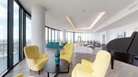 This elegant high-rise amenity space is designed to impress, offering an open, single-floor layout bathed in natural light from sweeping floor-to-ceiling curved glass windows that frame breathtaking panoramic city skyline views. The interior is anchored by a crisp, all-white palette complemented by light hardwood flooring throughout, creating a bright and airy atmosphere.

The expansive lounge area is thoughtfully divided into multiple seating zones, including clusters of vibrant yellow velvet armchairs arranged around round coffee tables, a teal sectional sofa seating area positioned directly against the curved window wall to maximize views, and a more formal lounge zone featuring plush white sectional sofas and gray accent chairs. Tray ceilings with warm recessed LED cove lighting add architectural depth and a refined glow to the space.

A standout feature is the grand black piano, positioned as a focal point alongside curated contemporary artwork displayed on wainscoted white walls, flanked by gold wall sconces. A flat-screen television is also integrated into this area, making it functional for events and gatherings.

The formal dining area accommodates a large party at a sleek dark dining table with modern black-frame chairs, illuminated by a striking cluster of geometric gold pendant lights. White cabinetry lines the back wall, completing this polished and versatile entertaining space.