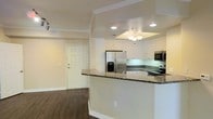 This single-floor space showcases a thoughtfully designed open-concept layout that seamlessly connects the kitchen and living areas. The kitchen is a standout feature, highlighted by crisp white cabinetry that extends to the ceiling, dark granite countertops with contrasting light veining, and a full suite of stainless steel appliances, including a French door refrigerator, microwave, and range. A raised peninsula counter serves as a natural divider between the kitchen and the adjacent living space, offering additional seating or prep space.

Crown molding frames the rooms with a refined touch, while a tray ceiling above the kitchen is accented by a elegant chandelier, adding a warm focal point to the space. Recessed lighting provides consistent, practical illumination throughout. The continuous dark hardwood flooring grounds the space and creates a rich contrast against the light-colored walls and white trim. A paneled entry door with classic hardware completes the welcoming entryway. The overall aesthetic is clean, bright, and move-in ready, combining functional design with modern, upscale finishes.