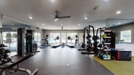 The Quad fitness center is a thoughtfully designed, single-level gym space that balances cardio, strength, and functional training in one cohesive layout. The interior is finished with neutral gray walls, rubber flooring throughout, and recessed lighting complemented by dual ceiling fans to maintain airflow and comfort during workouts.

The cardio zone is positioned along the windowed perimeter wall, where multiple treadmills and elliptical trainers benefit from abundant natural light and direct views of an outdoor pool area. Glass doors in this section provide easy access to the poolside amenity space, reinforcing the connection between indoor and outdoor recreation.

The strength training area features a substantial power rack with an Olympic barbell and weight plates, a full dumbbell rack with a flat bench, and a multi-function cable machine. A large wall mirror in this zone enhances the sense of space and provides functional feedback during lifts.

A dedicated functional training zone is set apart with yellow-green flooring tiles and includes a punching bag, resistance accessories, and wall-mounted training equipment. A bold, graphic accent wall branded with "The Quad" adds an energetic, motivational atmosphere to this corner.

Overall, the fitness center offers a comprehensive training environment with a modern aesthetic, ample natural light, and clear pool sightlines throughout.