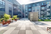 Cyber Deck is a thoughtfully designed outdoor amenity courtyard located in Everett, MA, nestled within a modern multi-story residential building. The space is enclosed by the building's contemporary facade, characterized by gray panel siding, large windows, and private balconies that overlook the courtyard below.

The courtyard is anchored by a generous turf lawn area, which serves as a central gathering hub. A large outdoor projection screen mounted on a repurposed shipping container makes the lawn ideal for open-air movie screenings. Adirondack chairs and woven pouf ottomans are arranged across the lawn to create a relaxed, social atmosphere.

A standout architectural feature is the converted shipping container, which doubles as a private meeting or lounge space. Inside, a stone-top table and woven chairs provide a functional and unique setting for small gatherings or work sessions.

The surrounding patio is finished with large-format checkerboard-patterned pavers and is thoughtfully divided into distinct seating zones. One area features upholstered sofas and lounge chairs arranged around fire features, while another offers dining tables and colorful chairs suited for casual meals or social gatherings. Lush planters with ferns and greenery are integrated throughout, adding a natural, refreshing contrast to the modern hardscape.