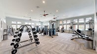 Located at 2300 SW 38th Ave in Ocala, Florida, this professional-grade fitness center spans nearly 2,000 square feet across two exercise rooms, offering a comprehensive and well-appointed workout environment. The main exercise floor is generously proportioned, featuring soaring ceilings, elegant herringbone-patterned luxury vinyl flooring, and a bank of large windows along the perimeter that flood the space with natural light and offer views of the surrounding outdoor areas. Ceiling fans are suspended throughout, enhancing airflow and comfort during workouts. The main floor is outfitted with an extensive array of strength and cardio equipment, including cable machines, selectorized weight stations, treadmills, ellipticals, rowing machines, a full dumbbell rack, kettlebells, medicine balls, adjustable benches, and plyometric boxes. Wall-mounted televisions and full-length mirrors enhance both functionality and the sense of spaciousness. A dedicated secondary room serves as a cycling and functional training studio, featuring stationary bikes and air bikes arranged before a continuous floor-to-ceiling mirror wall, a recessed tray ceiling with warm ambient lighting, and a wall-mounted display screen. The two rooms are connected via a sleek glass door, creating a cohesive and modern fitness facility.