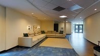 Located at 646 W Bay Street in the heart of Savannah, Georgia, this single-floor space offers a thoughtfully designed interior that balances comfort and functionality. The main lounge area serves as the focal point of the space, featuring a large, neutral-toned sectional sofa accented with decorative pillows, arranged atop a light area rug set against patterned blue-gray carpeting. Wall-mounted sconce lighting flanks a world map artwork, adding a touch of character and warmth to the neutral walls. The drop-panel ceiling incorporates recessed and flush lighting fixtures that ensure a well-lit, inviting atmosphere throughout. The open floor plan connects seamlessly to adjacent areas, with a curved architectural detail on one side adding a dynamic visual element. Toward the rear of the space, large glass entry doors allow natural light to filter in and provide a view of the surrounding greenery, enhancing the bright and airy feel. The overall ambiance is clean, modern, and professional, making it well-suited for a variety of commercial or hospitality uses. The thoughtful arrangement of furniture and lighting creates a space that feels both expansive and welcoming to visitors.