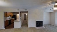 This single-floor residence offers a straightforward, functional layout centered around an open living area. The main living space is finished with wall-to-wall carpet and features a built-in fireplace framed by a clean white surround, serving as a focal point along one wall. Ceiling vents indicate a central HVAC system throughout.

Adjacent to the living area, the kitchen is partially open to the main space via a breakfast bar counter, providing a natural division while maintaining visual connectivity. The kitchen features wooden cabinetry, a contrasting dark refrigerator, and lighter countertops, with tile or hard flooring underfoot distinct from the carpeted living area.

A central hallway extends from the main living space, leading toward additional rooms at the rear of the residence. The hallway provides access to what appears to be a closet or utility area, with a door visible at the far end. Throughout the space, white walls and a textured ceiling create a neutral, cohesive backdrop. Overhead ventilation registers are positioned throughout, ensuring consistent climate control across the layout.