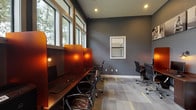 This single-floor business center is designed to provide a functional and professional workspace environment. The space is characterized by its bold, dark charcoal-gray accent walls, which create a focused and modern atmosphere. Patterned modular carpet tiles in gray, white, and yellow accent tones cover the floors throughout, adding a contemporary visual element underfoot.

The primary workstation area features a long, dark wood-finished desk running along one wall, equipped with multiple computer monitors, individual task lighting with warm-toned Edison-style bulbs, and semi-private divider panels in a rich amber hue, offering each user a degree of privacy. Ergonomic black office chairs are positioned at each station, accommodating several users simultaneously.

A generous row of large windows along one wall floods the workspace with abundant natural light and offers views of surrounding greenery, creating an open and refreshing ambiance. Additional workstations are arranged in a separate area of the room, providing flexible seating arrangements.

Elevated ceilings enhance the sense of openness, while recessed lighting supplements the natural light throughout the space. Black-and-white canvas artwork is displayed on the upper portion of one wall, adding a refined aesthetic touch. A glass-paneled door provides access to an adjacent area, contributing to the overall connectivity and flow of the space.