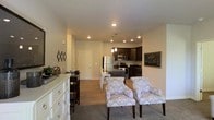 This single-floor space showcases an open-concept layout that seamlessly connects the living, dining, and kitchen areas into one cohesive and functional environment. The living area is anchored by plush patterned accent chairs and soft carpeting, creating a comfortable and inviting atmosphere. A white dresser with chrome hardware adds a clean, classic touch to the space.

The transition from carpet to hardwood-style flooring marks the shift into the dining and kitchen zones. A compact dining area with bar-height seating sits adjacent to the kitchen, making it ideal for casual meals. The kitchen is well-appointed with dark espresso cabinetry, stainless steel appliances including a refrigerator, microwave, and range, and ample counter space. Pendant lighting above the kitchen area adds a stylish focal point.

Recessed lighting throughout ensures the space remains bright and evenly lit. Neutral beige walls and light-toned ceilings contribute to an airy, open feel. A chalkboard-style wall feature near the kitchen adds a functional and decorative element. The overall design balances modern aesthetics with everyday practicality.