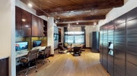 This single-floor space blends a warm, sophisticated aesthetic with practical functionality. The most striking architectural feature is the richly grained wood ceiling with exposed beams arranged in a herringbone pattern, lending the interior a refined, rustic character. Track lighting and a circular pendant fixture illuminate the space, complementing the natural light that filters in through multiple windows dressed with slatted blinds.

The layout is thoughtfully divided into two distinct zones. On one side, a built-in workstation with dark wood cabinetry, open shelving, and an integrated desktop computer provides a focused environment for productivity. The cabinetry extends floor to ceiling, offering substantial storage. On the opposite side, a casual lounge area features low-profile chairs and a coffee table arranged around a central gathering space, ideal for informal meetings or relaxed conversation.

Light hardwood flooring runs throughout, contrasting pleasantly with the darker tones of the ceiling and cabinetry. A large metal locker unit anchors one wall, adding an industrial element to the otherwise warm palette. An accent wall with a geometric patterned wallpaper introduces a contemporary visual detail. Overall, the space conveys a polished, professional ambiance with inviting warmth.
