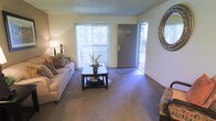 This single-floor unit centers around a thoughtfully arranged living area that serves as the heart of the space. The room is finished with wall-to-wall carpeting that adds warmth underfoot, while light-toned walls create a bright and neutral backdrop throughout. A large sliding glass door fitted with vertical blinds allows generous natural light to filter into the room and provides a view of the outdoor greenery beyond, enhancing the overall sense of openness.

The entry area, accessible through a solid front door, transitions seamlessly into the main living space, where there is a clear delineation between the carpeted interior and a hard-surface entryway. A decorative round mirror mounted near the entry adds a stylish accent and contributes to the room's welcoming atmosphere.

The living area comfortably accommodates a sofa and additional seating arranged around a dark wood coffee table with a lower storage shelf, offering both functionality and visual grounding to the space. A floor lamp provides supplemental ambient lighting. The textured ceiling adds a subtle architectural layer, and the overall layout is practical and well-proportioned, lending itself to comfortable everyday living.