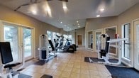 This fitness center offers a comprehensive workout environment designed to accommodate a variety of exercise needs. The space is laid out on a single floor, with light beige tile flooring throughout that contributes to a clean, open feel. Recessed lighting in the vaulted ceiling provides bright, even illumination across the entire room, complemented by an abundance of natural light streaming in through large windows along the walls.

The cardio section is well-appointed with multiple elliptical machines and treadmills arranged in a row, allowing users to exercise while enjoying views of the outdoors. Strength training equipment, including cable and weight machines, is positioned on both sides of the room, offering a balanced and functional layout. Rubber floor mats are placed beneath the equipment to ensure safety and protect the flooring.

Mirrored panels along one wall enhance the sense of space while allowing users to monitor their form during workouts. Frosted glass French doors lead to an adjoining area, adding architectural interest and further expanding the usability of the facility. The neutral gray walls and vaulted ceiling lend the space a modern, airy ambiance, making it an inviting and practical destination for fitness enthusiasts.