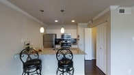 This single-floor residence showcases a thoughtfully designed interior that balances functionality with modern style. The kitchen serves as a central highlight, featuring granite countertops with warm, neutral toning that complement the crisp white cabinetry. Stainless steel appliances, including a refrigerator, range, and microwave, lend a contemporary feel, while a decorative dark tile backsplash adds visual contrast and texture behind the cooking area. A peninsula-style breakfast bar extends the counter space and is paired with elegant wrought iron bar stools, creating a casual dining and gathering spot. Overhead, two pendant lights with frosted glass shades hang at staggered heights, providing both task lighting and an aesthetic focal point. Recessed lighting throughout ensures the space is well-lit. Crown molding along the ceiling adds a refined architectural detail, elevating the overall finish of the interior. Rich dark hardwood flooring grounds the space and flows seamlessly through the visible areas, offering a striking contrast to the white cabinetry and light-toned walls. Interior doors are classic panel-style in white, maintaining a cohesive and clean design language throughout the home.