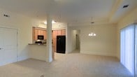 This single-floor space offers an open-concept layout that seamlessly connects the living area and kitchen, creating a cohesive and functional environment. The living area is finished with plush, light-toned carpeting that adds warmth and comfort underfoot, while a decorative chandelier hangs overhead, providing both ambient lighting and a refined focal point. Crown molding along the upper walls adds an architectural detail that elevates the overall aesthetic.

The kitchen is thoughtfully designed with light wood cabinetry that provides generous storage space. Dark granite countertops offer a striking contrast, while a raised breakfast bar creates a natural divide between the kitchen and living area, ideal for casual dining or entertaining. Black appliances, including a side-by-side refrigerator and built-in microwave, complement the kitchen's contemporary style. Overhead fluorescent lighting ensures the kitchen is well-lit for meal preparation.

A structural column near the kitchen entrance adds architectural character to the open floor plan. A hallway visible from the living area suggests additional rooms beyond, contributing to the overall sense of depth and livability. The neutral color palette throughout — soft whites and warm beiges — creates a bright, welcoming atmosphere well-suited for a variety of interior styles.