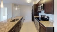 This single-floor unit is thoughtfully designed with a clean, neutral palette of warm beige tones and crisp white trim throughout. The open living area features rich hardwood-style flooring and is illuminated by track lighting, creating a bright and welcoming atmosphere. A doorway connects the living space to the carpeted bedroom areas, ensuring a comfortable flow between rooms.

The bedrooms are generously sized and finished with plush carpeting. Large floor-to-ceiling windows flood the rooms with abundant natural light while offering expansive outdoor views of the surrounding cityscape. Vertical blinds provide privacy and light control.

The kitchen is well-appointed with dark espresso cabinetry, granite countertops, tile flooring, and a full suite of stainless steel appliances, including a refrigerator, electric range, microwave, and dishwasher. A pendant light adds a stylish accent above the sink.

Two full bathrooms feature tile flooring, dark vanity cabinetry with white countertops, and tub-shower combinations with white subway tile surrounds. One bathroom provides direct access to a walk-in closet, adding convenient functionality to the primary suite.