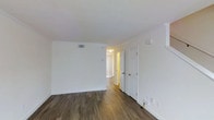 This two-bedroom, two-bathroom residence spans two floors, offering a functional and well-organized layout. The interior is finished with rich, dark wood-style plank flooring that runs throughout, providing a warm contrast to the crisp white walls and white trim. The open living area is bright and clean, with smooth ceilings, recessed ventilation, and ample wall space. A vertical blind-covered window allows natural light to filter into the space, creating an inviting atmosphere. A hallway connects the living area to additional rooms, with a paneled white door visible to the right, hinting at thoughtfully separated living spaces. The neutral color palette throughout provides a versatile backdrop, making the space easy to furnish and personalize. The two-floor layout offers added separation between living and private areas, a practical feature for residents seeking distinct functional zones. Overall, the unit presents a tidy, move-in-ready environment with consistent finishes and a straightforward design that prioritizes comfort and usability.
