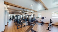 Located on Clinton Keith Road in Murrieta, CA, this well-appointed fitness center offers an expansive, single-floor exercise space designed to support a wide range of workout routines. The room's architecture is defined by decorative exposed wood beams set against a clean white ceiling, lending a modern yet warm aesthetic throughout. Large ceiling fans ensure comfortable airflow, while recessed lighting and multiple windows along the walls flood the space with natural light and provide views of the surrounding outdoor landscape.

The flooring is finished in a durable hardwood-style material that extends seamlessly across the entire area, providing both a polished appearance and a practical surface for exercise. The strength training zone is well-equipped with a comprehensive dumbbell rack, weight benches, and multiple cable and cable-crossover machines. A large arched wall mirror adds depth to the space while supporting form-check during workouts. Wall-mounted flat-screen televisions are positioned throughout for entertainment.

The cardio section features treadmills, stationary cycling bikes, and rowing machines, all equipped with individual digital screens. Exercise mats offer dedicated space for stretching and floor work. A tiled water station with an arched architectural surround adds a refined finishing touch. Overall, the space presents a clean, functional, and inviting environment for fitness enthusiasts.