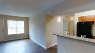 This single-floor residence offers a thoughtfully arranged open layout that connects the living and kitchen areas seamlessly. The living space features rich, dark hardwood flooring that extends throughout, grounding the interior with warmth and character. Neutral, light-toned walls create a bright and welcoming ambiance, complemented by natural light filtering in through a window fitted with blinds.

A defining feature of the space is the kitchen bar, which provides a natural transition between the cooking and living areas. The bar is topped with granite countertops displaying an elegant stone pattern, adding a touch of sophistication to the overall design. The kitchen area also includes a wall-mounted light fixture that adds focused illumination to the workspace.

Beyond the kitchen bar, an open doorway leads to an additional room, suggesting a practical and well-connected floor plan. White baseboards throughout the space provide clean, crisp contrast against the hardwood floors and painted walls, contributing to a polished, move-in-ready aesthetic.

Overall, the space presents a functional and inviting environment with a balance of practical design elements and tasteful finishes, making it well-suited for comfortable everyday living.