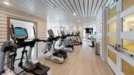 This fitness center is thoughtfully designed across multiple levels connected by a sleek staircase with contemporary cable railings featuring wood-topped handrails. The upper level is dedicated to cardio training, housing a row of True 400 treadmills and elliptical machines aligned along large windows that invite ample natural light. Full-length mirrors and wall-mounted flat-screen televisions enhance the space, while light hardwood-style flooring runs throughout, contributing to a clean, modern aesthetic. Recessed lighting and slatted wood ceiling panels add architectural character to portions of the space.

The lower level accommodates strength training equipment, providing a dedicated area for resistance-based workouts. A separate studio space features floor-to-ceiling mirrors, a wall-mounted television, and cycling bikes, making it suitable for spin sessions or group fitness. Motivational fitness photography adorns the walls throughout, reinforcing the space's active atmosphere. The overall design balances functionality with a polished, contemporary aesthetic, creating an inviting and well-organized environment for a comprehensive workout experience.