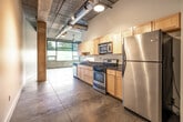 This single-floor loft-style unit showcases a distinctive urban industrial aesthetic throughout its well-organized layout. Exposed concrete ceilings with visible ductwork and structural concrete columns create a raw, warehouse-inspired character, while large-format tile and polished concrete flooring provide a clean, durable foundation underfoot. Pendant lighting adds warm accents against the industrial backdrop.

The open living area benefits from natural light streaming through large windows that frame outdoor greenery, lending an airy quality to the space. The galley-style kitchen is thoughtfully equipped with light maple cabinetry, dark granite countertops, stainless steel appliances including a gas range, microwave, dishwasher, and refrigerator — offering full functionality within a compact footprint.

The bathroom features a tiled tub-and-shower combination with striking dark slate wall tile, a light wood vanity with a dark countertop and stainless steel vessel sink, and a wall-mounted sconce, creating a modern, polished look. An in-unit washer and dryer area adds practical convenience. The overall ambiance of the unit blends industrial rawness with clean, modern finishes, making it a distinctive and functional urban living space.