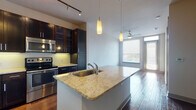 This well-appointed single-floor residence showcases a thoughtfully designed open-concept layout that seamlessly connects the kitchen, dining, and living areas. The kitchen is a standout feature, boasting rich espresso-toned cabinetry that extends to the ceiling for generous storage, complemented by light granite countertops, a tile backsplash, and a full suite of stainless steel appliances including a refrigerator, range, and built-in microwave. A substantial central island with a granite surface and integrated sink serves as both a functional workspace and a natural gathering point, illuminated by elegant pendant lights overhead. Track lighting along the ceiling provides additional task lighting throughout the kitchen.

Rich hardwood flooring flows through the main living and kitchen areas, adding warmth and continuity to the space. The living area benefits from large windows dressed with horizontal blinds, inviting plentiful natural light and featuring a ceiling fan for comfort. A carpeted bedroom offers a quieter retreat, with its own window providing soft, diffused natural light.

The bathroom is finished to a high standard, featuring large-format white tile flooring, a dark-framed vanity with granite countertops, a well-lit mirror, and a glass-enclosed shower. The overall aesthetic is contemporary and cohesive, with consistent use of dark cabinetry, granite surfaces, and modern fixtures throughout.