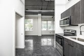 Located at 600 E Fifth St in Des Moines, Iowa, this one-bedroom, one-bathroom unit delivers an industrial-chic aesthetic with a thoughtfully modern interior. Polished concrete floors extend throughout the entire space, reflecting natural light and anchoring the unit's contemporary character. Exposed ductwork and structural beams along the ceiling reinforce the industrial design language while adding visual depth and height to the open living area.

The living space is bright and open, benefiting from multiple large windows fitted with vertical blinds that invite generous natural light and views of the surrounding neighborhood. The kitchen is efficiently designed with dark wood-grain cabinetry, white quartz countertops, a matte black industrial-style faucet, and a full suite of stainless steel appliances including a range, over-the-range microwave, dishwasher, and refrigerator. A bold steel entry door adds an industrial accent.

The generously sized primary bedroom offers polished concrete flooring, a large window, and ample closet storage with built-in shelving. The bathroom is a standout, featuring large-format dark slate-style wall tiles in the walk-in shower, a crisp white vanity with a white countertop, and matte black fixtures and hardware throughout, creating a striking monochromatic contrast that ties the space together cohesively.