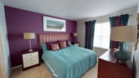 This single-floor space showcases a thoughtfully designed bedroom that serves as its standout feature. The room is anchored by a bold purple accent wall, creating a rich and vibrant focal point that contrasts elegantly with the neutral tones of the surrounding walls and light beige carpeting. A generously sized bed, dressed in soft teal bedding and adorned with layered decorative pillows in coordinating purples and stripes, occupies the center of the room and immediately draws the eye. A striped upholstered headboard ties together the room's color palette with effortless cohesion. Flanking the bed are matching nightstands topped with warm table lamps, adding symmetry and a cozy ambiance to the space. A large window dressed with floor-length teal and burgundy curtains allows natural light to filter in, brightening the room while maintaining a sense of warmth and intimacy. A framed landscape artwork above the headboard adds an artistic touch, enhancing the room's carefully curated aesthetic. The overall atmosphere is inviting and comfortable, with a bold yet harmonious color scheme that lends the space a distinctive and polished character.