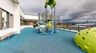 Located at 2453 Renfrew St in Vancouver, BC, this rooftop outdoor play area offers a thoughtfully designed environment for children, elevated above the surrounding neighbourhood with panoramic views of the Vancouver skyline and beyond. The expansive rooftop terrace is finished with a vibrant teal rubber safety surface that provides both visual appeal and a cushioned, durable play area underfoot.

At the heart of the space stands a multi-level playground structure featuring climbing frames, a spiral slide, a yellow dome climbing igloo, and various interactive components rendered in bright lime green and teal tones. Natural-form seating elements — including log-style stools and a simple bench — are positioned along the quieter zones of the terrace, offering a more relaxed area for observation or rest.

The building facade bordering the rooftop incorporates a contemporary design with white panel cladding, warm horizontal wood accents, and large windows that look out onto the play area. A glass and steel perimeter railing encloses the terrace, ensuring safety while preserving unobstructed sightlines to the cityscape, including the downtown Vancouver skyline visible in the distance. The space benefits from open-sky exposure, bringing abundant natural light throughout the day and creating an airy, open ambiance in an urban rooftop setting.