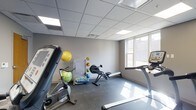 The fitness room offers a practical and well-organized space designed to support a variety of workout routines. The room features a drop tile ceiling with recessed fluorescent lighting panels that provide bright, even illumination throughout. A large window along one wall allows natural light to fill the space, creating an open and energizing atmosphere.

The flooring is a durable, smooth concrete-style surface well-suited for fitness activities. The room is equipped with a range of cardio machines, including a stationary cycling bike, a treadmill, and a rowing machine, offering users multiple options for cardiovascular training. Along the wall, a dedicated storage area holds an assortment of exercise balls in varying sizes, including medicine balls and stability balls, alongside other small fitness accessories. A folding mat or board is also visible, adding further versatility to the available equipment.

The neutral grey walls and clean, uncluttered layout contribute to a functional and focused environment. Overall, the fitness room provides a compact yet comprehensive space for residents or users to maintain an active lifestyle with a well-rounded selection of exercise options.