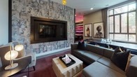 This thoughtfully designed single-floor lounge space is divided into several distinct zones, each with its own character while maintaining a cohesive contemporary aesthetic. The media lounge area anchors one end of the space, featuring a large wall-mounted flat-screen television framed by striking collage-style wallpaper depicting illustrated portraits. A plush sectional sofa, a light wood coffee table, and a pair of upholstered wingback chairs with accent pillows create a comfortable and inviting seating arrangement. Rich, deep-toned flooring adds warmth and contrast throughout this zone, while a sculptural cluster pendant light overhead adds artistic flair.

Transitioning further into the space, the layout opens into a more versatile social area with large-format tile flooring in a cool gray tone. This zone incorporates dark wood booth-style banquette seating upholstered in deep emerald green, paired with a round white pedestal table and cylindrical ottomans. Curated framed artwork adorns the walls, contributing to the gallery-like atmosphere. Adjacent to this, a counter-height bar seating area bathed in warm pink ambient lighting features sleek industrial-style stools. A backlit bar display is visible in the background, reinforcing the sophisticated, lounge-inspired character of the overall space.