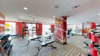 This fitness center delivers a dynamic and energizing workout environment designed to accommodate a wide range of exercise needs. The space is defined by its bold visual identity, featuring striking red accent walls and a decoratively painted structural column adorned with colorful circular artwork, creating an upbeat and motivating atmosphere throughout.

The layout is generously proportioned, with a smooth flow between dedicated strength and cardio zones. The strength training area includes adjustable weight benches, a multi-function cable and rack system, and an assortment of medicine balls and free weights neatly organized along the red accent wall. The cardio section is equipped with a row of elliptical trainers and upright exercise bikes, positioned to take advantage of the large windows that allow ample natural light to fill the space and offer exterior views.

Underfoot, a patterned multi-tone carpet tile adds texture and visual interest while defining the workout areas. The ceiling features recessed lighting and a ceiling fan, ensuring both bright illumination and comfortable airflow during exercise. Full-length mirrors along one wall enhance the sense of depth and allow users to monitor their form. Overall, this fitness center combines functional equipment with a lively, contemporary design aesthetic.
