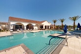 Located at 20807 W Indian School Rd in Buckeye, Arizona, this single-story community amenity center showcases a striking Spanish Colonial Revival architectural style, characterized by crisp white stucco exterior walls, decorative arched colonnades, and warm terracotta clay tile roofing. The centerpiece of the outdoor space is a generously sized swimming pool with crystal-clear turquoise water, complete with accessible entry handrails and clearly marked depth indicators for safety. Surrounding the pool, a wide expanse of light-colored tile decking provides ample space for relaxation and recreation. The sun deck is furnished with modern aluminum chaise lounges outfitted with blue cushions and shaded by navy market umbrellas, creating a comfortable outdoor retreat. Towering desert palm trees are thoughtfully positioned throughout the space, adding lush vertical interest and a resort-like atmosphere. The clubhouse building features large, dark-framed windows that allow natural light to flow into the interior while offering pool views. Restroom and utility access points are conveniently integrated into the building's exterior. The overall ambiance evokes a luxurious desert resort, perfectly suited to the sunny Sonoran Desert climate of the greater Phoenix West Valley area.