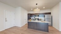 This thoughtfully designed single-floor residence offers a seamless open-concept layout that integrates the kitchen and living room into one cohesive, light-filled space. The kitchen serves as a centerpiece, featuring a large granite-topped island with an integrated sink, dark espresso cabinetry extending to the ceiling, a white subway tile backsplash, stainless steel appliances including a refrigerator, microwave, and range, and a stylish drum pendant light overhead. Recessed lighting and light wood-look flooring carry throughout the space, creating a clean, modern aesthetic.

The living area is open and airy, with multiple windows fitted with blinds that invite natural light, a glass-paneled entry door, and a neutral color palette that lends versatility to the space. A hallway connects the main living area to the private bedroom, which is generously sized, features a ceiling fan, a window with natural light, and the same consistent light wood flooring. A dedicated closet provides ample storage. The bathroom is efficiently designed with dark wood cabinetry, a granite vanity countertop, a large mirror, and light flooring that coordinates with the rest of the unit. A separate hallway enhances the flow and privacy of the layout.