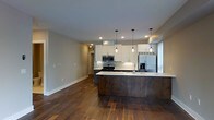 This single-floor space showcases a thoughtfully designed open-concept layout that seamlessly connects the kitchen and living areas. The kitchen is a standout feature, outfitted with crisp white upper cabinetry, dark lower cabinetry, and a classic subway tile backsplash that adds texture and visual interest. Stainless steel appliances, including a refrigerator with ice maker, gas range, and built-in microwave, provide a sleek, modern finish. A generously sized kitchen island with a clean white countertop serves as a natural focal point, offering ample prep space and casual seating potential. Three stylish pendant lights hang above the island, casting warm, focused light and adding a decorative touch. Recessed lighting throughout ensures the space remains bright and welcoming. Rich, wide-plank hardwood flooring flows throughout the open area, lending warmth and character to the contemporary design. The neutral gray wall tones create a versatile backdrop that complements the contrast between the light and dark cabinetry. Natural light filters in through a window near the refrigerator, enhancing the airy feel of the space. The overall ambiance is modern, clean, and functional.