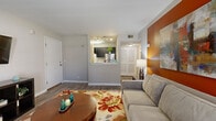 This single-floor residence offers a thoughtfully designed layout that balances comfort and functionality. The living area serves as the heart of the home, featuring durable wood-look flooring, neutral gray walls accented by a warm orange feature wall, and a cozy arrangement centered around a plush sofa and a round wood coffee table. A vibrant floral area rug adds a pop of color and texture underfoot.

The kitchen is accessible via a pass-through window from the living area, creating an open and connected feel. It is outfitted with dark cabinetry, light quartz-style countertops, a stainless steel double-basin sink, a dishwasher, and a range. White subway tile backsplash adds a clean, modern touch, while under-cabinet lighting enhances visibility and ambiance.

The bedroom is a serene retreat, carpeted for warmth and comfort, and featuring a large window dressed with blinds that allow natural light to filter in. Ample storage is provided by a dedicated closet with shelving, while the room offers enough space for a full furniture arrangement including a dresser with mirror.

Throughout the space, neutral tones create a calm, cohesive atmosphere, while carefully placed accent lighting and curated décor contribute to an inviting and livable environment.