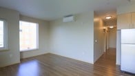 This single-floor space offers a clean, contemporary design highlighted by smooth light wood flooring that runs throughout the open layout. The main living area is characterized by crisp, neutral-toned walls that create a bright and airy atmosphere, complemented by a large window that allows natural light to fill the room and provides views of the surrounding exterior. A wall-mounted mini-split air conditioning unit ensures year-round climate control directly within the living space. The open floor plan provides a generous sense of space, with a clear sightline leading toward a hallway that connects to additional areas of the unit. The minimalist aesthetic is consistent throughout, with clean baseboards, subtle electrical outlets, and an absence of unnecessary ornamentation, lending the space a modern and functional character. The hallway beyond suggests additional rooms, maintaining the same neutral palette and light-toned finishes seen in the main area. Overall, the space presents a well-maintained, move-in-ready environment with a straightforward and practical layout suited to a variety of uses.