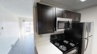 This single-floor space offers a thoughtfully designed layout that balances functionality and modern style. The kitchen serves as a focal point, showcasing rich, dark espresso-toned cabinetry with sleek hardware that creates a bold contrast against the light countertops and white walls. A stainless steel over-the-range microwave and a black electric coil range provide practical, full-featured cooking amenities, while the upper cabinetry offers generous storage. The kitchen opens directly into the living area, creating a connected, flowing floor plan that enhances the sense of space. The living area benefits from abundant natural light streaming through a large window, lending the space an airy, welcoming ambiance. Light-toned flooring throughout further brightens the interior and ties the rooms together cohesively. The drop ceiling with a textured finish adds a classic architectural element consistent throughout the space. Overall, the layout is practical and efficient, designed to maximize usability while maintaining a clean, contemporary aesthetic that feels both comfortable and inviting.