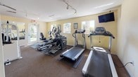 This fitness center is a fully equipped, single-floor facility designed to support a well-rounded exercise regimen. The space is finished with warm yellow walls, wood wainscoting accents, and durable carpet flooring throughout, creating a clean and comfortable workout environment.

The strength training area is well-appointed with multiple white cable machines, including a multi-functional cable crossover system and a leg press/extension machine, all arranged with ample space between equipment for safe and comfortable use. Full-length mirrors line one wall, allowing users to monitor their form during exercises and contributing to a sense of openness throughout the space.

The cardio section is equally comprehensive, featuring a lineup of treadmills, elliptical trainers, and a recumbent bike — all positioned near windows that frame outdoor greenery, bringing in natural light and pleasant views. Track lighting runs along the ceiling to ensure bright, even illumination throughout. A wall-mounted television is positioned within view of the cardio equipment for added convenience during workouts.

Additional amenities include a wall clock, posted informational signage, and a dedicated entry area with storage. An exterior door in the cardio zone provides convenient direct access. The overall layout is thoughtfully organized, balancing dedicated strength and cardio zones while maintaining an open, airy atmosphere.