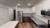 This single-floor unit showcases a thoughtfully designed layout with modern finishes and a clean, contemporary aesthetic throughout. The kitchen is a standout feature, outfitted with stainless steel appliances including a full-size refrigerator, electric range with over-the-range microwave, and dishwasher. Two-tone cabinetry pairs crisp white upper cabinets with dark espresso lower cabinets, complemented by a classic subway tile backsplash and dark countertops. A central island with a contrasting navy base provides additional prep and storage space, while durable wood-look flooring ties the kitchen and open living area together seamlessly. The living area is bright and open, benefiting from a large bay window that frames lush outdoor greenery, allowing generous natural light to fill the space. The bedroom features plush carpeting, a ceiling fan, and dual windows that offer additional natural light and outdoor views. The bathroom is finished with large-format dark tile on both the shower surround and floor, a soaking tub, and a floating dark-wood vanity with white countertop, creating a polished, spa-like atmosphere. Recessed lighting throughout the unit adds to the clean, modern feel of the overall space.
