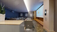 This private screening room, located in the 94597 area of California, is a thoughtfully designed cinematic space that combines high-end audiovisual technology with refined interior design. The room is organized across a single floor, offering a generously proportioned viewing environment with tiered seating rows that ensure unobstructed sightlines to a large wall-mounted projection screen framed by warm natural wood cabinetry and built-in speakers below.

Seating consists of rows of plush, neutral-toned lounge chairs arranged in a stadium-style configuration, separated by a low wooden divider rail that adds to the architectural definition of the space. The ceiling features indirect cove lighting that casts a soft, ambient glow complementing recessed downlights. Deep navy-blue accent walls contrast with warm wood tones and taupe finishes, creating an immersive yet sophisticated atmosphere.

A dedicated entryway greets visitors with a sculptural 3D-textured wall panel backlit for visual impact, built-in cabinetry with a dark stone countertop, and a full-height glass-paneled door. Framed artwork is displayed throughout, adding a gallery-like quality. Textured wall-to-wall carpeting completes the space, enhancing acoustics while reinforcing the room's premium aesthetic.