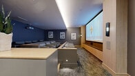 This private screening room, located in the 94597 area of California, is a thoughtfully designed cinematic space that combines high-end audiovisual technology with refined interior design. The room is organized across a single floor, offering a generously proportioned viewing environment with tiered seating rows that ensure unobstructed sightlines to a large wall-mounted projection screen framed by warm natural wood cabinetry and built-in speakers below.

Seating consists of rows of plush, neutral-toned lounge chairs arranged in a stadium-style configuration, separated by a low wooden divider rail that adds to the architectural definition of the space. The ceiling features indirect cove lighting that casts a soft, ambient glow complementing recessed downlights. Deep navy-blue accent walls contrast with warm wood tones and taupe finishes, creating an immersive yet sophisticated atmosphere.

A dedicated entryway greets visitors with a sculptural 3D-textured wall panel backlit for visual impact, built-in cabinetry with a dark stone countertop, and a full-height glass-paneled door. Framed artwork is displayed throughout, adding a gallery-like quality. Textured wall-to-wall carpeting completes the space, enhancing acoustics while reinforcing the room's premium aesthetic.