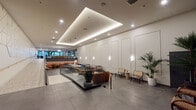Located on the iconic Wilshire Boulevard in Los Angeles, this single-floor commercial space offers a polished and thoughtfully designed interior that blends contemporary aesthetics with warm, inviting touches. The main lobby area is defined by a striking coffered ceiling with integrated LED lighting that casts a soft, ambient glow across the expansive grey stone tile flooring. Panels of crisp white wainscoting line the walls, complemented by sleek wall-mounted sconces that add dimension and warmth.

A glass-railed mezzanine-style seating platform anchors the central zone, furnished with a mix of rattan chairs, leather accent seating, and small side tables that invite gathering and collaboration. Lush tropical potted palms are placed throughout, adding organic contrast to the refined architectural palette. A lounge area deeper within the space features vibrant upholstered banquette seating in rust and gold tones, set against a textured geometric wall panel with pendant lighting above.

The elevator lobby presents a more dramatic character, with dark fluted stone cladding and warm metallic lift doors creating a bold, grounded aesthetic. Nearby, a quieter seating alcove with charcoal panel-molded walls offers a more intimate retreat.