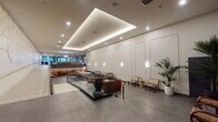 Located on the iconic Wilshire Boulevard in Los Angeles, this single-floor commercial space offers a polished and thoughtfully designed interior that blends contemporary aesthetics with warm, inviting touches. The main lobby area is defined by a striking coffered ceiling with integrated LED lighting that casts a soft, ambient glow across the expansive grey stone tile flooring. Panels of crisp white wainscoting line the walls, complemented by sleek wall-mounted sconces that add dimension and warmth.

A glass-railed mezzanine-style seating platform anchors the central zone, furnished with a mix of rattan chairs, leather accent seating, and small side tables that invite gathering and collaboration. Lush tropical potted palms are placed throughout, adding organic contrast to the refined architectural palette. A lounge area deeper within the space features vibrant upholstered banquette seating in rust and gold tones, set against a textured geometric wall panel with pendant lighting above.

The elevator lobby presents a more dramatic character, with dark fluted stone cladding and warm metallic lift doors creating a bold, grounded aesthetic. Nearby, a quieter seating alcove with charcoal panel-molded walls offers a more intimate retreat.