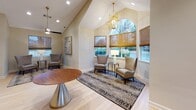 This well-appointed clubhouse offers a thoughtfully designed single-floor layout that blends comfort and sophistication. The main lounge area immediately impresses with its soaring vaulted ceilings, recessed lighting, and a decorative gold geometric chandelier that adds a refined touch. Light hardwood flooring runs throughout, complementing the warm, neutral wall tones and creating a bright, welcoming ambiance. Seating areas are arranged around patterned area rugs, featuring elegant wingback chairs and accent tables, ideal for casual gatherings or relaxation. Natural light flows generously through multiple windows dressed with bamboo roll-up shades, including a striking arched window that serves as a focal architectural feature. A decorative sunburst mirror and framed artwork adorn the walls, enhancing the space's curated aesthetic. A ceiling fan with a modern finish adds both function and style to the vaulted ceiling. The kitchen area is outfitted with bold, floor-to-ceiling dark navy cabinetry accented by gold hardware, white marble-look countertops, and a mirrored subway tile backsplash. Full-size appliances, including a refrigerator and microwave, make the kitchen fully functional for entertaining and events. The overall design strikes a balance between modern elegance and inviting warmth.